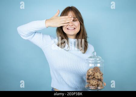 Junge schöne Frau hält Schokolade Chips Cookies Glas über isolierten blauen Hintergrund Augen mit Händen lächelnd fröhlich und lustig. Blinde Kontra Stockfoto