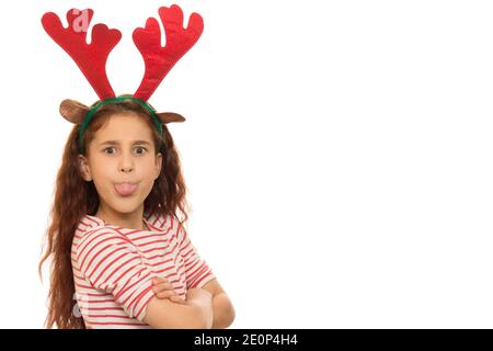 Liebenswert junge langhaarige Mädchen trägt Weihnachten Regenhirsch Geweihe kleben Ihre Zunge ist wütend auf die Kamera, die mit ihr posiert Arme isoliert gefaltet Stockfoto