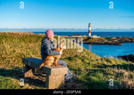 Ein Hundespaziergänger ruht sich auf einer Bank aus, die den rot-weißen Leuchtturm im Dorf Boddam bei Peterhead in Aberdeenshire, Schottland, Großbritannien, überblickt Stockfoto