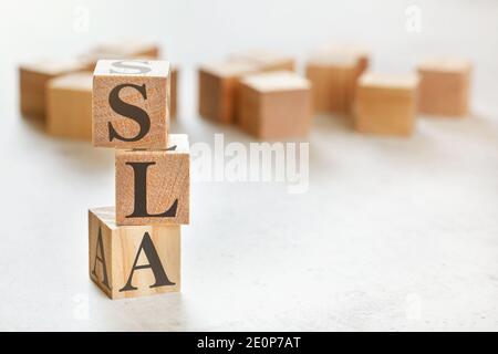 Drei Holzwürfel mit den Buchstaben SLA (bedeutet Service Level Agreement), auf weißem Tisch, mehr im Hintergrund, Platz für Text in der rechten unteren Ecke Stockfoto