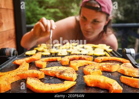 Butternut Squash Stücke auf elektrischen Grill gegrillt, konzentrieren sich auf helle orange Gemüse gewürzt mit Gewürzen, verschwommen junge Frau im Hintergrund Stockfoto