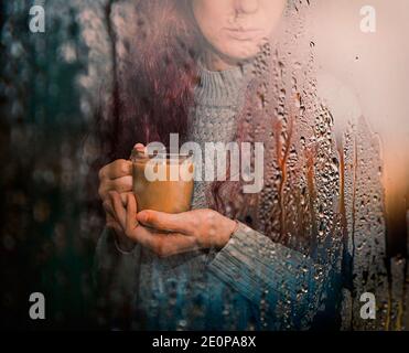 Mädchen mit einer Tasse Kaffee schaut auf den Regen Durch nasses Glas Stockfoto