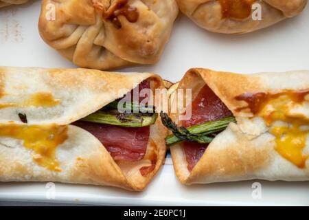 Makroansicht der Zenitale. Horizontale Ansicht von Säcken mit Käse und Tomaten gefüllt. Serrano Schinken und grüne Spargelbrötchen auf einem weißen Keramikblech Stockfoto