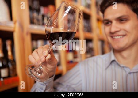 Nahaufnahme eines Rotweinglases in der Hand eines fröhlichen Mannes im Weinladen. Glücklicher männlicher Kunde, der Rotwein vor dem Kauf verkostet Stockfoto