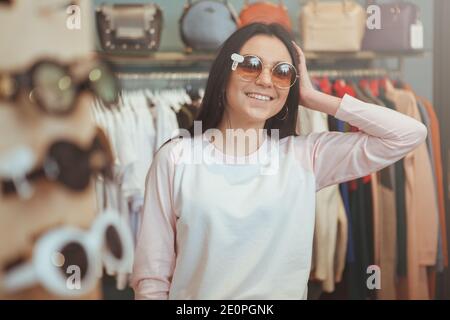 Glückliche junge schöne Frau lächelnd, Blick in den Spiegel, versuchen, neue Sonnenbrille im Modegeschäft. Schöne fröhliche weibliche Kunden genießen sh Stockfoto