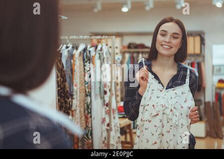 Glückliche junge schöne Frau lächelnd fröhlich versucht ein neues Kleid in Bekleidungsgeschäft vor dem Spiegel. Attraktive weibliche Kundin im Stockfoto