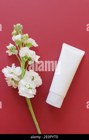 Blank weiße Squeeze Flasche Plastikschlauch auf einem burgunderrot / roten Hintergrund mit einer weißen Blume. Ansicht von oben, Platz für einen Text. Vertikales Foto. Stockfoto