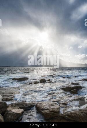 Sonnenstrahlen platzen durch Sturmwolken über Calvi im Balagne Region von Korsika mit der felsigen Mittelmeerküste in Der Vordergrund Stockfoto