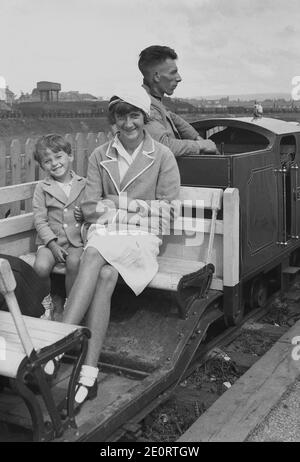 1940er Jahre, historisch, ein süß lächelnder, glücklicher Junge mit seiner großen Schwester in ihrem Schulblazer, der zusammen auf einer Holzbank vor einem Zug von dem Fahrer auf einer Miniatureisenbahn sitzt und auf eine Fahrt wartet, England, Großbritannien. Stockfoto