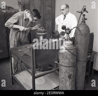 1950er Jahre, historisch, unter dem wachsamen Auge eines weiß beschichteten Tutors, eines jungen Auszubildenden oder Lehrlings in der Luftfahrttechnik an einer Werkbank, mit einem großen Blasbrenner, um zwei Metalle zusammen zu löten, eine kreisförmige Metallplatte auf einem quadratischen Metallsockel in Short Bros, Rochester, England, Großbritannien. Stockfoto