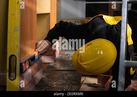 Beim Verlegen der dekorativen Ziegel muss man mit der Wasserwaage von Zeit zu Zeit prüfen, ob die vertikalen und horizontalen richtig sind. Ein Mitarbeiter, der persönliche Schutzausrüstung trägt. Stockfoto