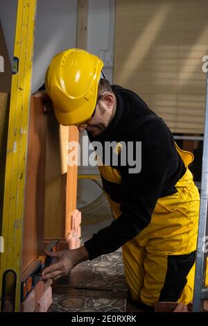 Beim Verlegen der dekorativen Ziegel muss man mit der Wasserwaage von Zeit zu Zeit prüfen, ob die vertikalen und horizontalen richtig sind. Ein Mitarbeiter, der persönliche Schutzausrüstung trägt. Stockfoto