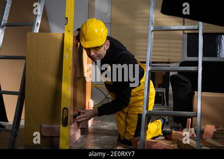 Beim Verlegen der dekorativen Ziegel muss man mit der Wasserwaage von Zeit zu Zeit prüfen, ob die vertikalen und horizontalen richtig sind. Ein Mitarbeiter, der persönliche Schutzausrüstung trägt. Stockfoto