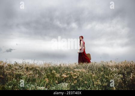 Ein einsames rotes Haar Mädchen in roten Kleid hält ein Vintage Koffer und Wandern in der Natur Stockfoto