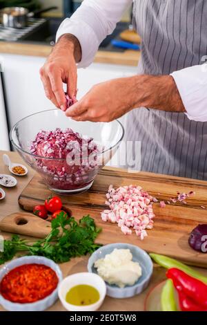 Der Koch fügt die Zwiebel in eine transparente Glasschüssel mit Fleisch, während auf der Seite der Arbeitsfläche gibt es Gemüse, Scheiben Speck, Gewürze a Stockfoto