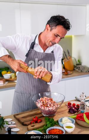 Der Koch fügt die Gewürze in eine transparente Glasschüssel mit Fleisch, Zwiebel und Speck, während auf der Seite der Arbeitsfläche gibt es Gemüse, Scheiben Stockfoto