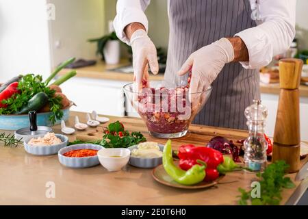 Der Koch Mischen Fleischbällchen mit Gewürzen und Gemüse in einer transparenten Glasschüssel, während auf der Seite der Arbeitsfläche gibt es Gemüse, Scheiben o Stockfoto