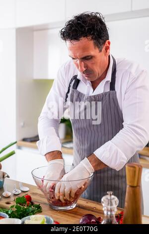 Der Koch Mischen Fleischbällchen mit Gewürzen und Gemüse in einer transparenten Glasschüssel, während auf der Seite der Arbeitsfläche gibt es Gemüse, Scheiben o Stockfoto