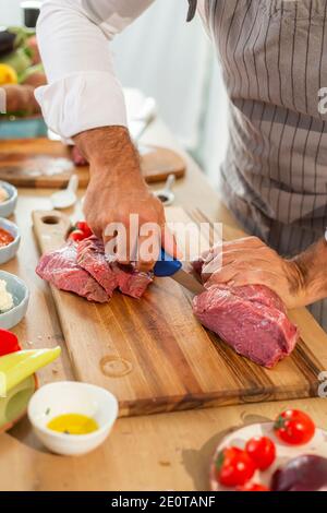 Eine Nahaufnahme der Hände des Küchenchefs, die Fleisch mit einem Messer in dünne Stücke schneiden. Verschiedene Gewürze und Gemüse können rund gesehen werden. Stockfoto