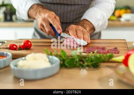 Eine Nahaufnahme der Hände des Küchenchefs, die während der Vorbereitungsphase Carpaccio-Fleisch auf einem Holzbrett in dünne Stücke schneiden. Verschiedene kleine Küche b Stockfoto