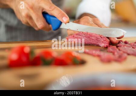 Eine Nahaufnahme der Hände des Küchenchefs, die während der Vorbereitungsphase Carpaccio-Fleisch auf einem Holzbrett in dünne Stücke schneiden. Verschiedene kleine Küche b Stockfoto