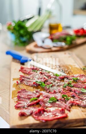 Nahaufnahme auf einem hölzernen Küchenbrett aus vorgefertigten Carpaccio-Fleisch zum Verkosten. Verschiedene Gemüse kann um gesehen werden, sowie kleine Küche b Stockfoto