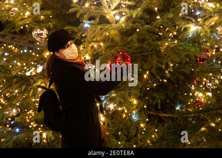 Frau hält Weihnachten riesige rote Kugel. Frau mit Maske steht vor dem großen Weihnachtsbaum. Blick auf die Kamera. Stockfoto