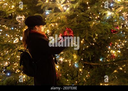 Frau hält Weihnachten riesige rote Kugel. Frau mit Maske steht vor dem großen Weihnachtsbaum. Stockfoto