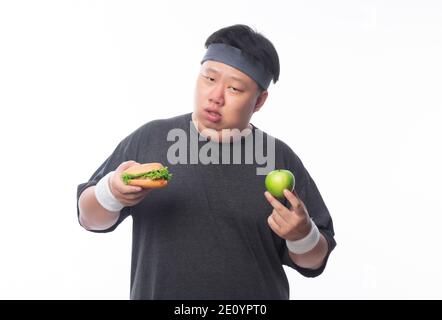 Junge asiatische lustige Fett Sportmann hält Hamburger und grünen Apfel isoliert auf weißem Hintergrund. Gesundes Lifestyle-Konzept. Stockfoto