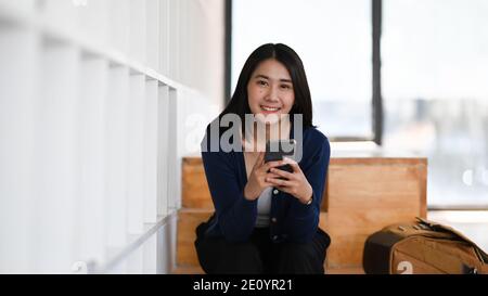 Fröhliche junge Frau Hände halten Smartphone und lächeln an der Kamera, während in der Bibliothek sitzen. Stockfoto