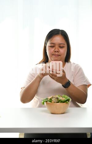 Portrait der übergewichtigen jungen Frau mit Smartphone Foto von Salat in einer Schüssel zu Hause. Stockfoto