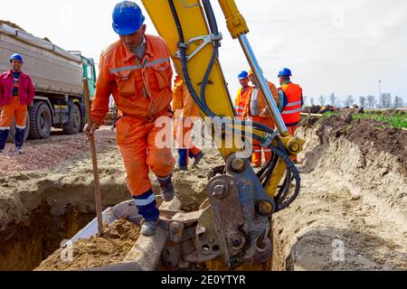 Arbeiter ist Sprung aus quadratischen Graben mit wenig Hilfe von Bagger Eimer voll von ausgegrabenen Boden. Stockfoto