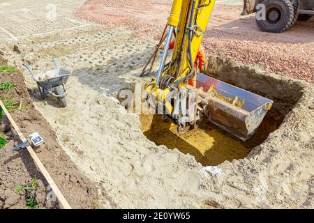 Arbeiter ist Sprung aus quadratischen Graben mit wenig Hilfe von Bagger Eimer voll von ausgegrabenen Boden. Stockfoto