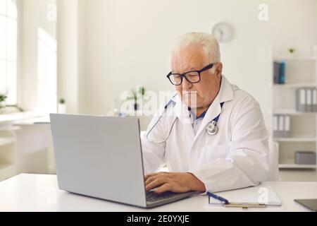 Serious Senior Doctor Tippen auf Laptop-Tastatur sitzen am Schreibtisch Im Klinikbüro Stockfoto