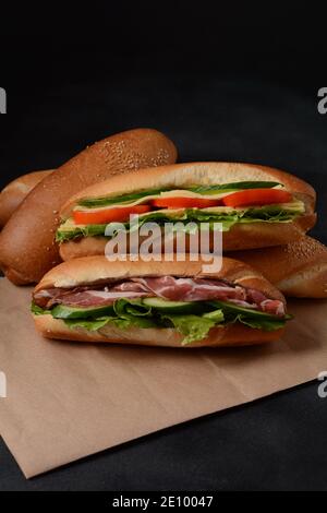 Frische U-Boot-Sandwiches mit Speck, Käse, Tomaten, Salat, Gurken und Zwiebeln auf dunklem Hintergrund Stockfoto