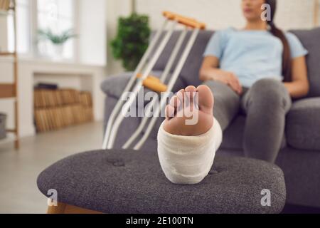 Frau mit Bein in Gips gegossen sitzt auf dem Sofa an Zuhause mit zwei Krücken daneben Stockfoto