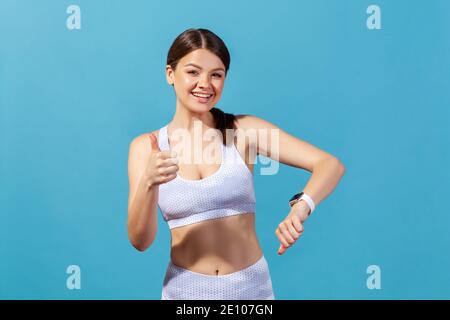 Glückliche athletische Frau in weißer Sportbekleidung zeigt Daumen nach oben demonstriert Smartwatch an ihrem Handgelenk, Anzeigen überprüfen, zufrieden mit Workout-Ergebnisse. Stockfoto