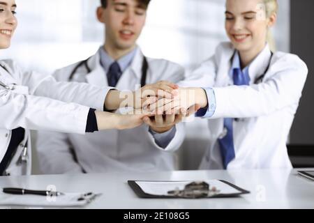 Gruppe von Ärzten, die sich als Kreis an den Händen anschlossen und bereit sind, Patienten zu helfen, während sie am Schreibtisch im modernen Krankenhausbüro sitzen. Erfolgreiche Teamarbeit in Stockfoto