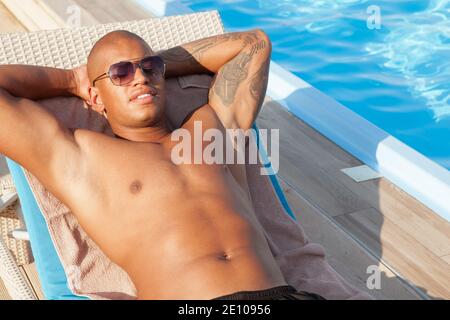 Kurzer Schuss eines hübschen afrikanischen Mannes mit Sonnenbrille, der am Pool in der Sonne liegt. Attraktiver Mann, der neben dem Swimmingpool bräunt Stockfoto