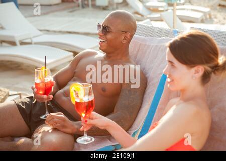 Glückliches junges Paar lachend, mit Cocktails am Strand in luxuriösen Badeort. Schöner afrikanischer Mann, der sich mit seiner Freundin am Strand entspannt Stockfoto