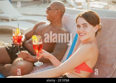 Glücklich schöne junge Frau ina Bikini ähnlich der Kamera, ihr schöner Freund Sonnenbaden auf dem Hintergrund. Multiethnisches Paar, das sich im Th Stockfoto