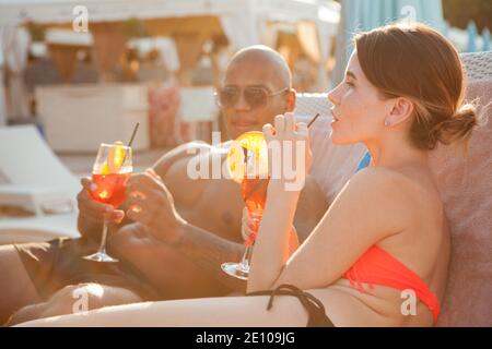 Schöne junge Frau genießt trinken Cocktail am Strand mit ihrem Freund. Multirassisches Paar, das am Strand zusammen relaxt Stockfoto