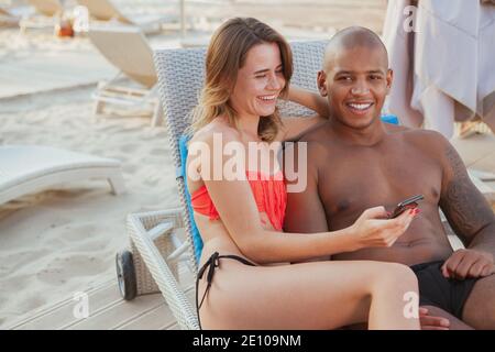 Glücklicher schöner afrikanischer Mann lachend, genießt am Strand mit seiner schönen Freundin liegen, kopieren Raum. Junge multiethnische Paare genießen Travelli Stockfoto