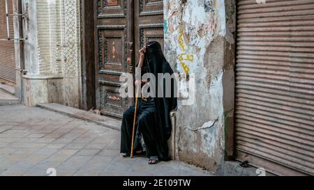 Yazd, Iran - Mai 2019: Alte iranische muslimische Frau in Tschador, das schwarze Kleid, bedeckt ihr Gesicht, sitzt in einem sehr alten Gebäude Stockfoto