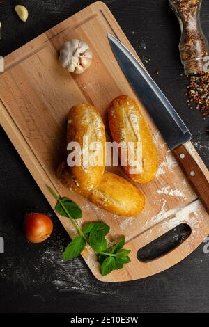 fresh wheat white bread on a cutting board with seasonings sprinkled with flour on a black wooden table Stock Photo