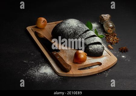 Frisches schwarzes Roggenbrot auf einem Schneidebrett mit Gewürzen Mit Mehl auf einem schwarzen Holztisch bestreut Stockfoto