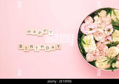 Happy Birthday Schriftzug auf den Holzquadraten mit Buchstaben auf dem rosa Hintergrund und großer runder Karton mit roten Blumen und Rosen. Grußkartenkonzept Stockfoto