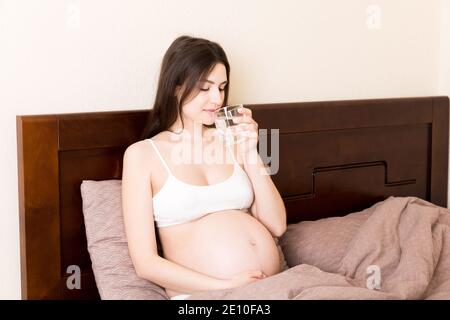 Foto von ziemlich schwangeren Frau halten ihre Hände auf dem Bauch, während Trinkwasser auf dem Bett zu Hause. Stockfoto