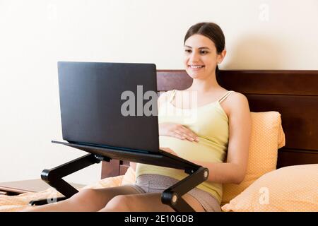 Schwangere Frau mit Laptop-Computer im Haus auf dem Bett. Schwangere Morgenarbeit Konzept. Stockfoto