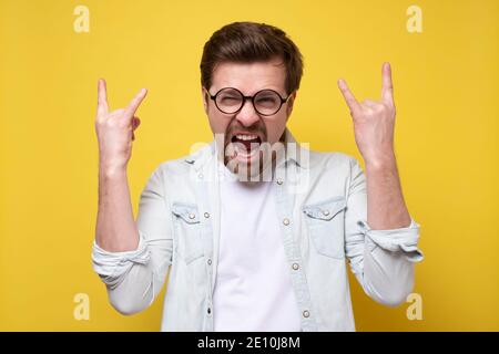 Kaukasischer Mann, der mit beiden Händen Felsschilder zeigt und laut schreit, Überwältigung und Freude ausdrückt. Studio auf gelber Wand. Stockfoto